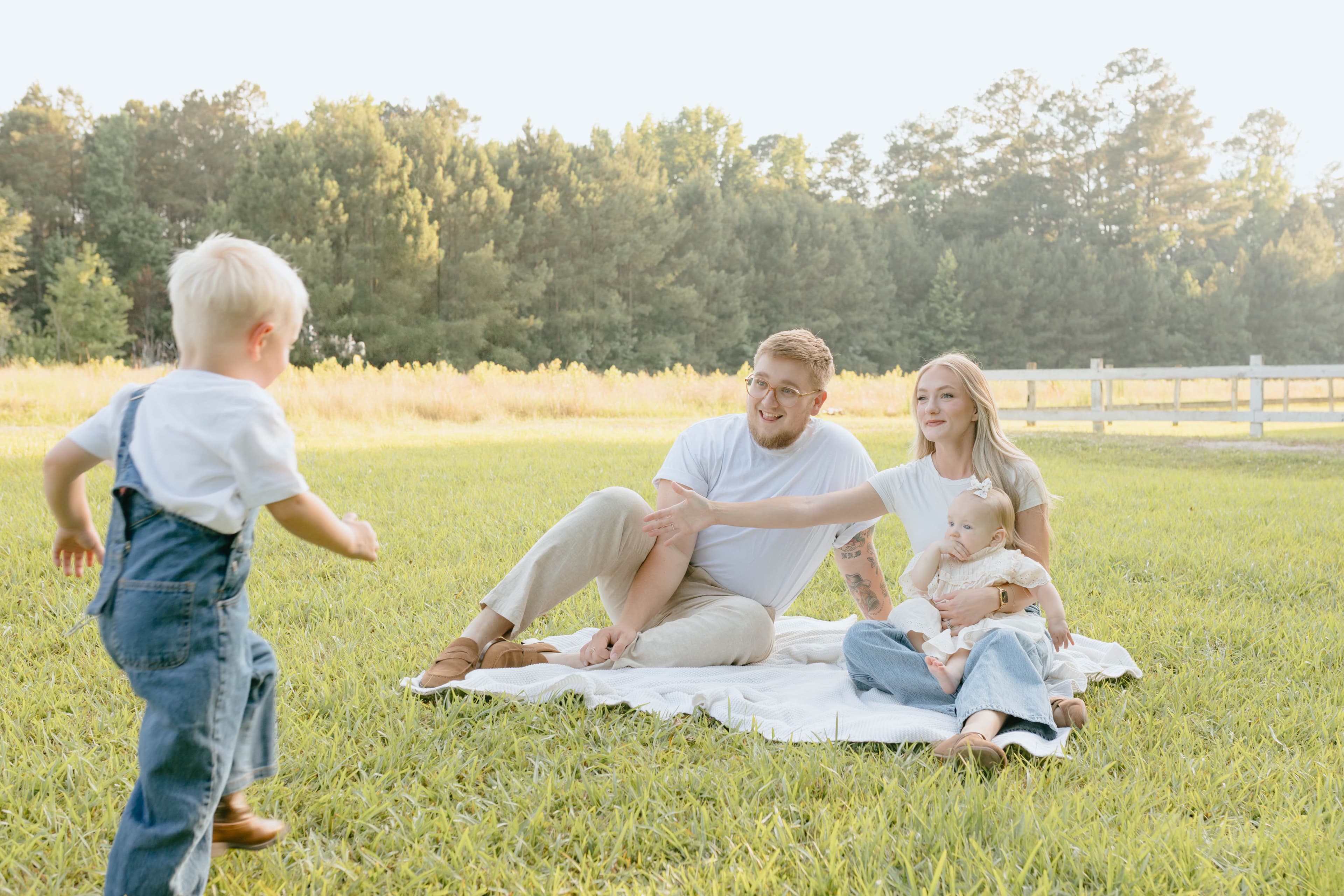 Boy in overalls runs toward his parents and baby sister sitting in a grassy field.