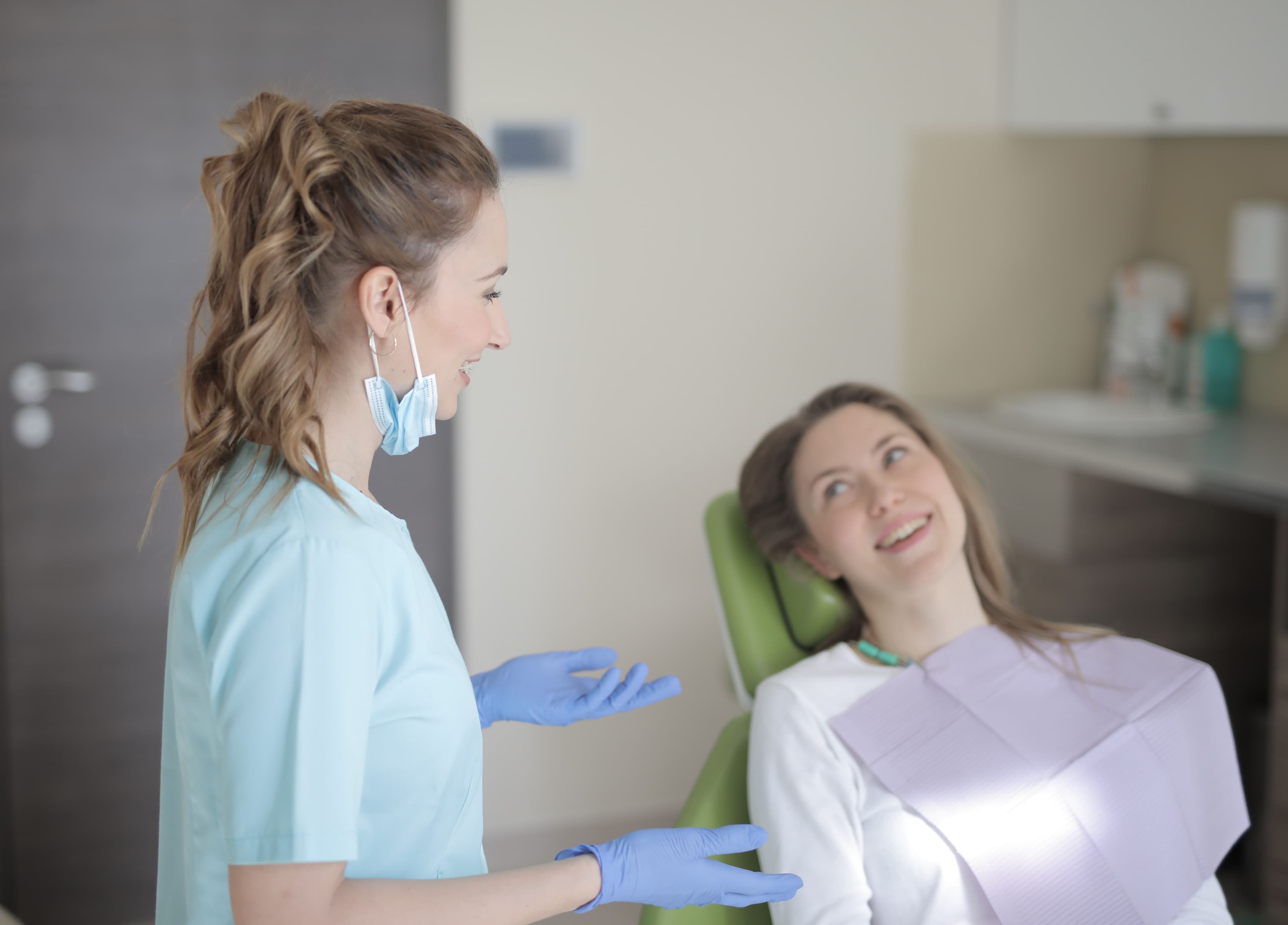 Smiling dentist in blue scrubs and gloves talks to a patient in a dental chair.